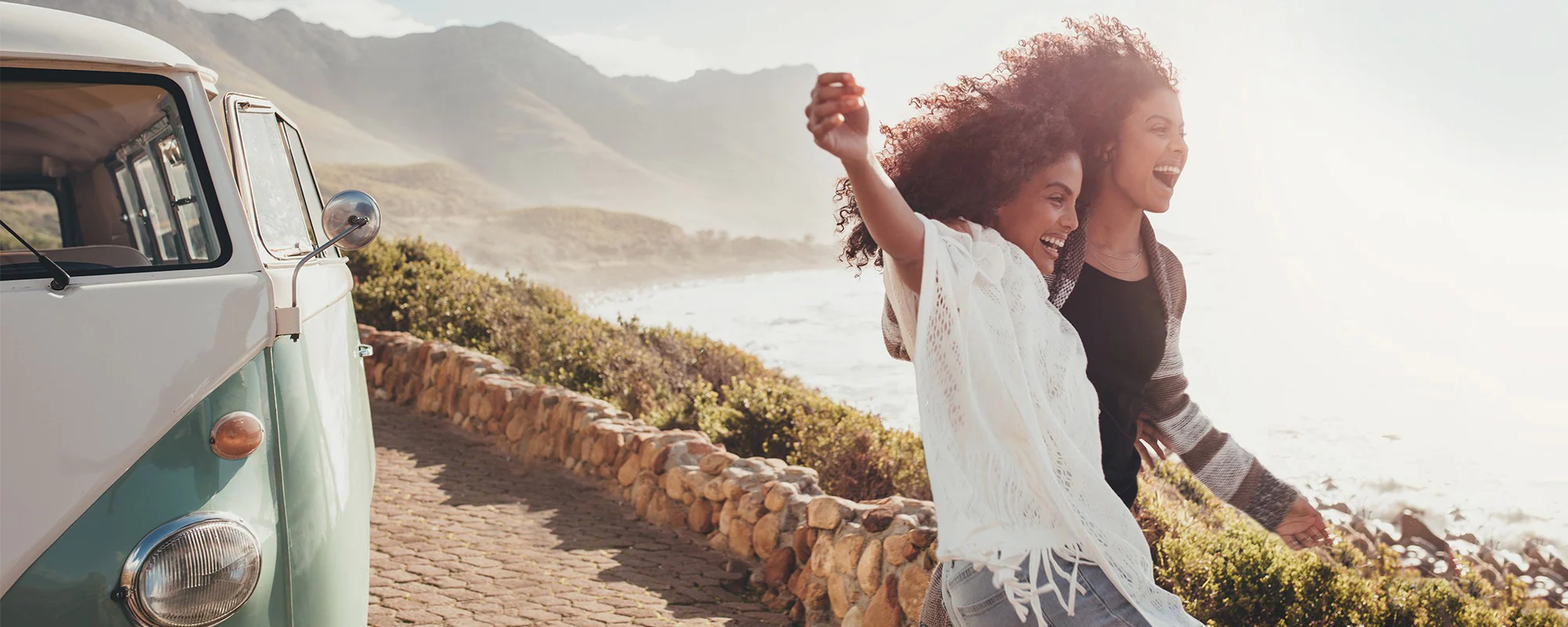 two people stand beside a van, embracing and looking out at the ocean, smiling